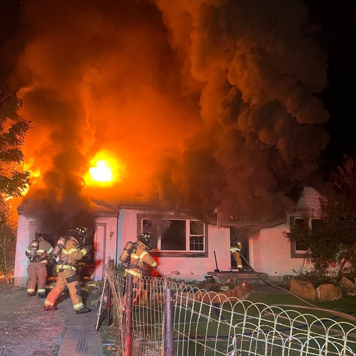Firefighters at night working a house fire with flames and heavy smoke from the roof